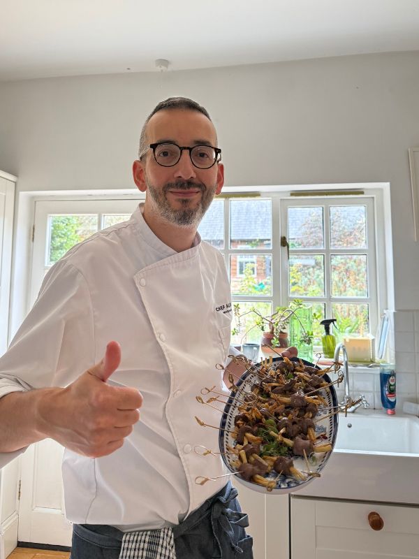 Chef Alpay holding a platter of skewered canapés and giving a thumbs-up in a home kitchen